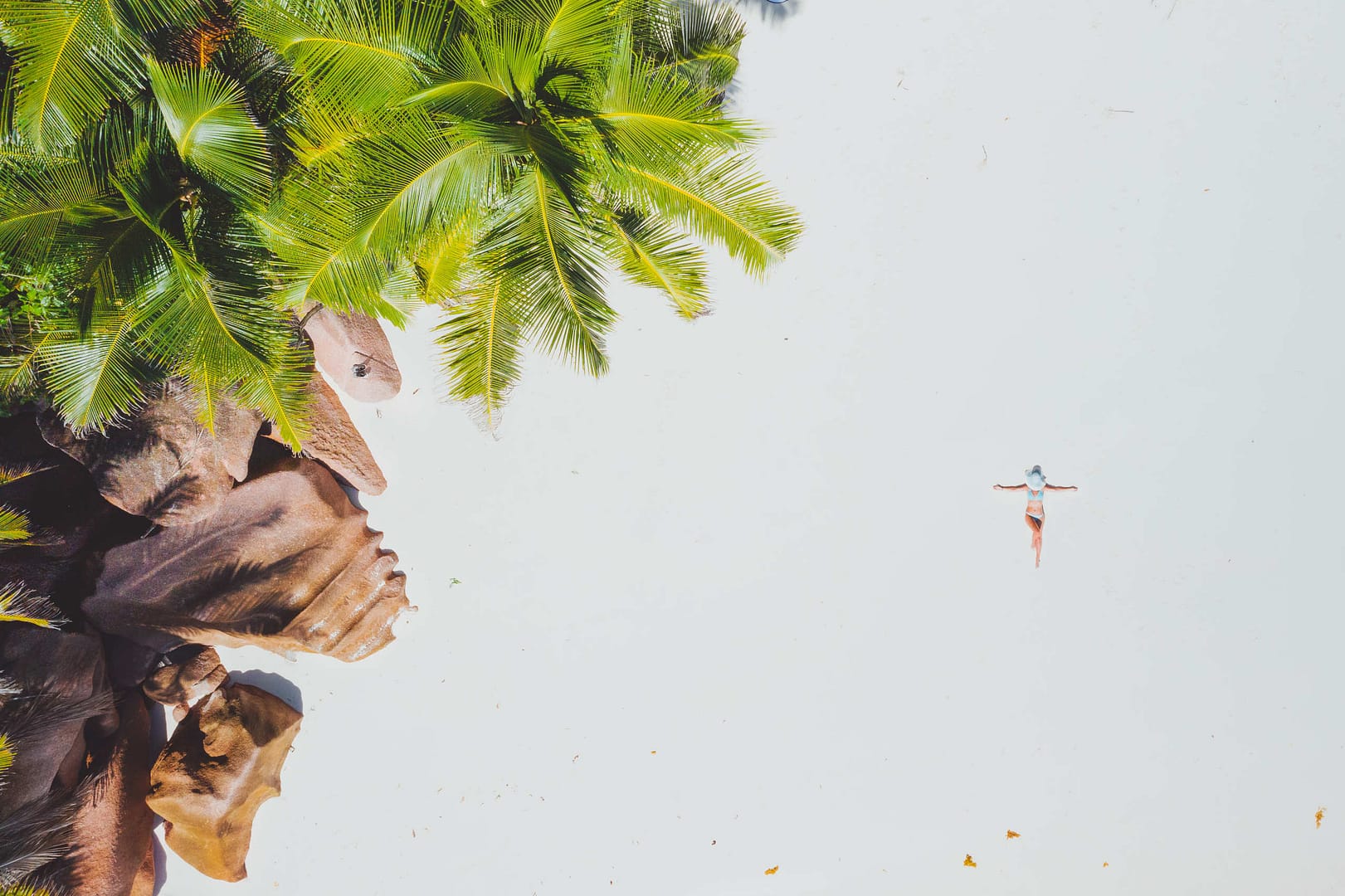 Aerial photo of young woman laying on exotic tropical beach with white sand. Girl sunbathing and relaxing near rockstone and palm trees. Concept of travel vacation holidays in paradise.